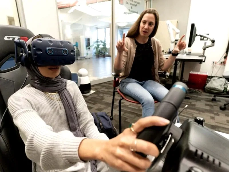 CMU student wearing a VR headset and using a driving simulator with a professor seated behind them instructing