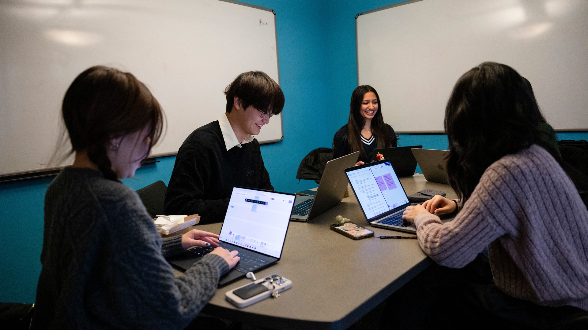Four students work on their laptops in a room with blue walls and whiteboards.