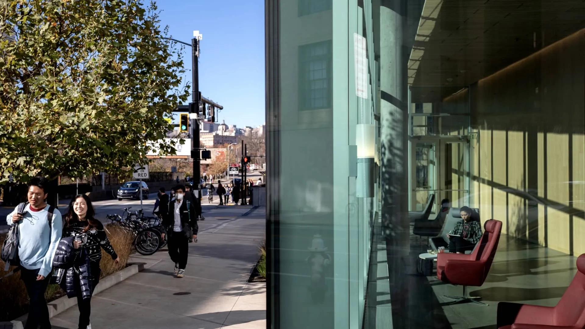 CMU students walking along a sunny sidewalk in front of a glass building with a CMU student seated in a chair working