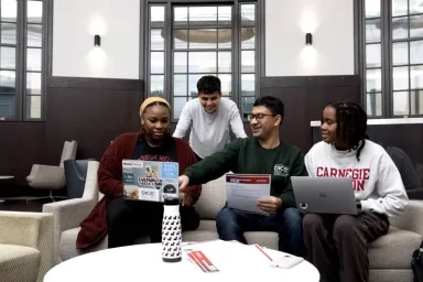 Four CMU students gathered on a couch, engaged in discussion with laptops and papers in a modern, well-lit campus lounge space